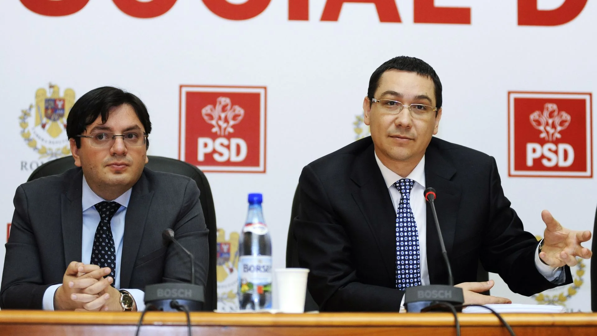 Doi oficiali stau la masa unei conferințe de presă, în fața siglelor PSD și ale Guvernului României.
