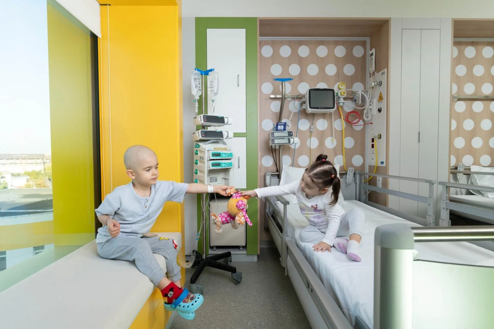 Doi copii într-un salon pediatric luminos, lângă un pat de spital și echipament medical.